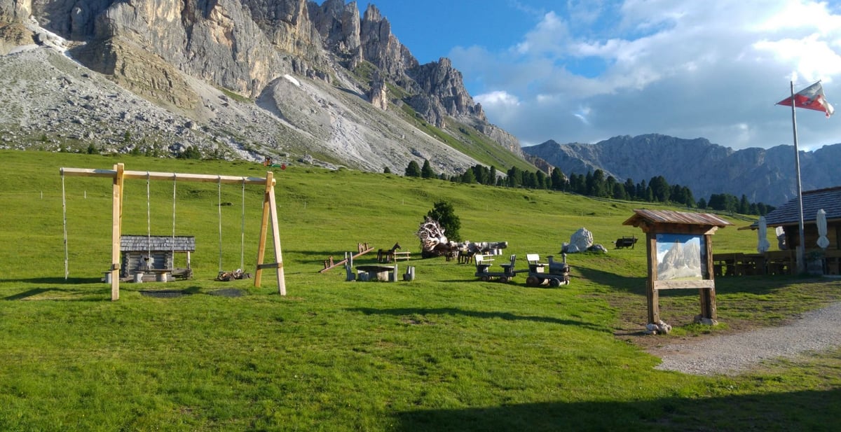 Alpine meadow with wooden swing set and playground equipment beneath dramatic mountain cliffs on a sunny day