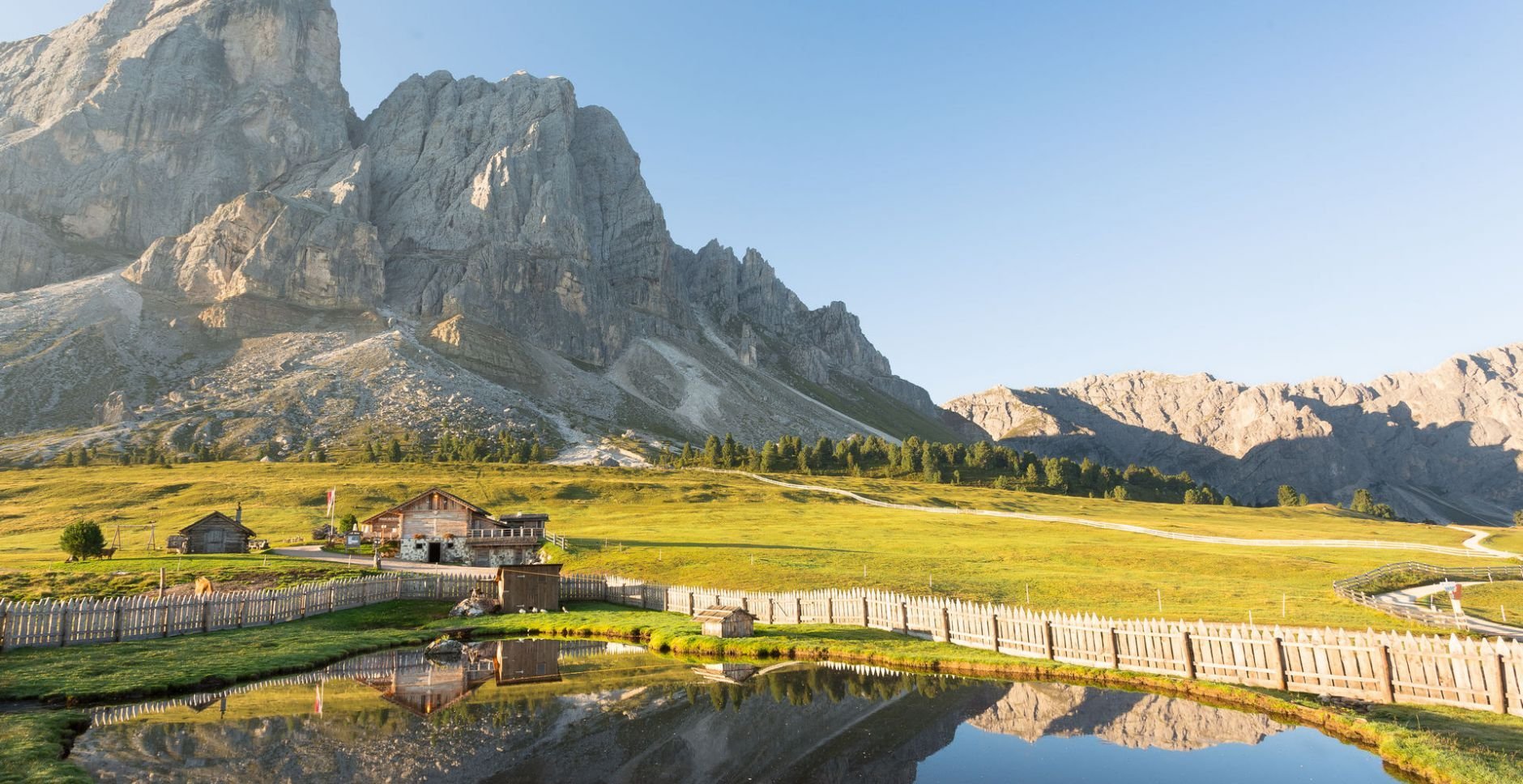 Alpine valley with wooden chalets, green meadows, and dramatic mountain peaks reflected in a small pond