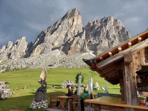 Wooden chapel with religious statues and cross overlooking green meadow and dramatic rocky mountain peaks