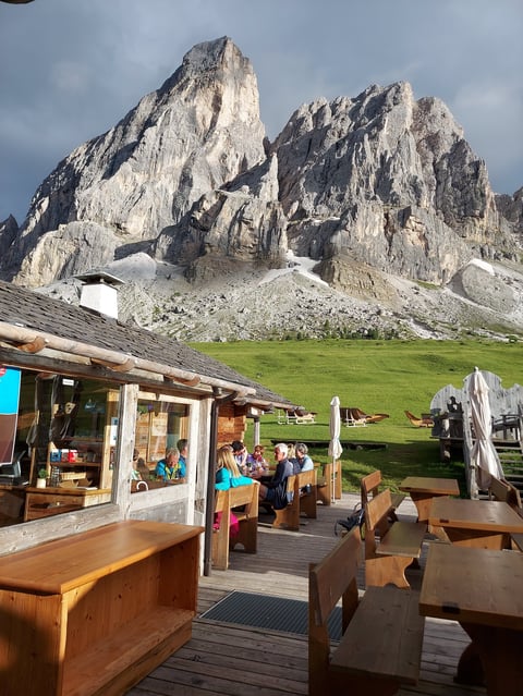 Mountain hut with wooden deck seating overlooking dramatic rocky peaks and green alpine meadow
