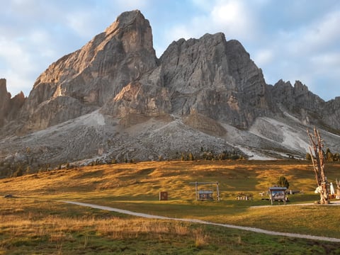 Dramatic rocky mountain peak with golden meadow, wooden structures, and cloudy sky in alpine landscape