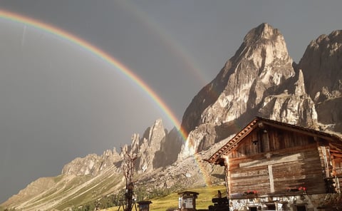 A wooden mountain hut nestled at the base of towering rocky peaks with a vibrant double rainbow arching overhead in the Alps