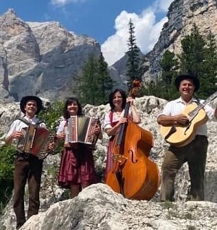 Five musicians in traditional Alpine clothing playing accordion, violin, and cello on rocky mountain terrain with snow-capped peaks in background